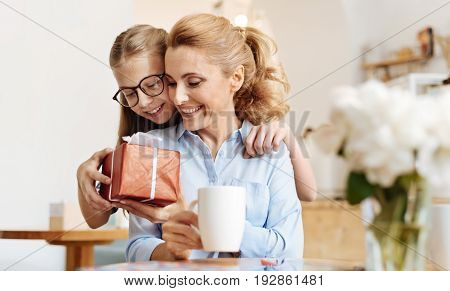 Love you mum. Winsome teenage girl hugging her dear mother from behind and giving her a beautifully wrapped gift for Mothers Day while the mother looking at it with affection