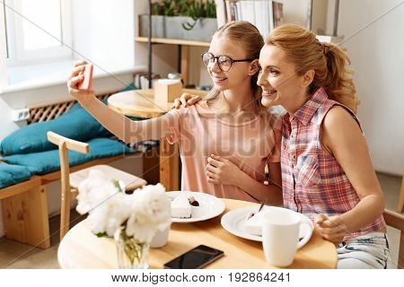 Happy faces. Joyous pretty girl and her elegant mother sitting at the cafe table, taking a selfie together and smiling at the camera broadly