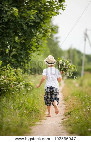 Back view on cute bare feet kid boy in straw hat running on a countryside road and holding bunch of wild flowers. Child running in the park. Outdoors. Happy summer and lifestyle concept. Childhood.