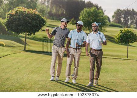 Confident Smiling Men In Caps And Sunglasses Holding Golf Clubs And Walking On Lawn
