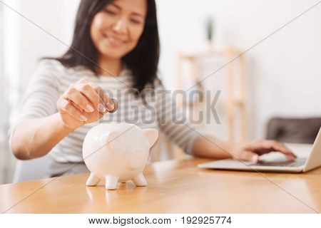 For future. Selective focus of a piggy bank standing on the table while cheerful asian woman putting coins inside and working on the laptop