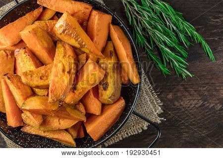 An overhead photo of roasted sweet potatoes in a pan, shot from above on dark rustic wooden textures with rosemary branches, with a place for text