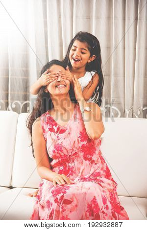 playful happy Indian little girl covering her mother's eyes and giving her surprise while sitting on white sofa in drawing room