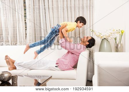 Happy Indian father playing with son while lying on sofa in living room at home. Asian Young man playing with small boy. child playfully punching his father or dad