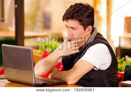 Handsome thoughtful young man at the bar working in his computer on a wooden table.