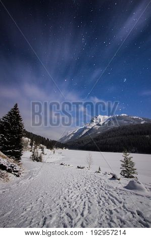 Frozen lake and snowy path in mountains during cold night under sky full of stars and faslty flowing clouds, Two Jack Lake, Banff, Canada