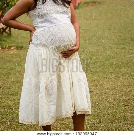Close up photo of young pregnant woman in elegant white stylish dress touching her belly with hands.