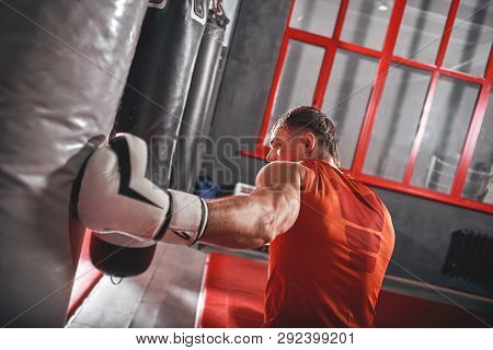 Strong Punch. Muscular Athlete In White Gloves Training On Heavy Punch Bag In Boxing Gym
