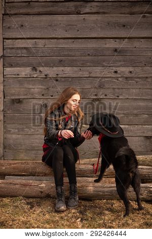 Happy Young Woman Plaing With Her Black Dog In Fron Of Old Wooden House. Girl Tries A Hat To Her Dog