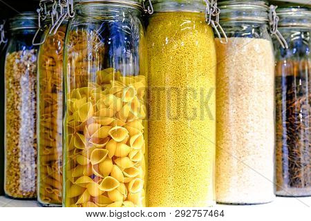Various Raw Cereals, Grains, Beans And Pasta For Healthy Cooking In Glass Jars On Kitchen Shelf, Whi