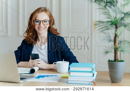 Photo Of Red Haired Smiling Young Woman Installs App On Mobile Phone, Checks Newsfeed, Works On Lapt