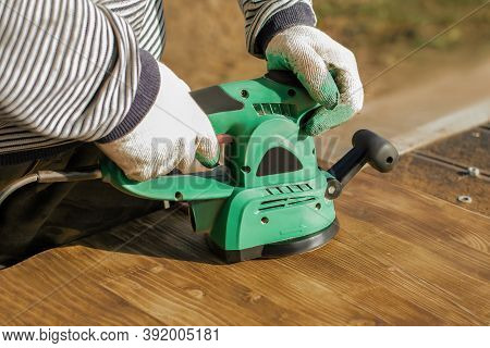 Carpenter In A Protective Gloves Is Grinding An Burnt Boards With An Electric Sander Outdoor. Home I