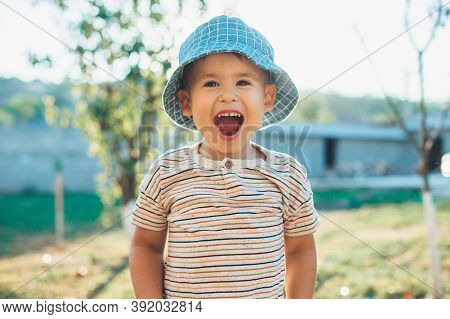 Happy Caucasian Boy In A Blue Hat Is Laughing At Camera While Playing In The Backyard Of The House