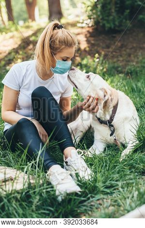 Blonde Caucasian Woman With Mask Is Enjoying Time Outside With Her Dog Lying On The Grass In A Park