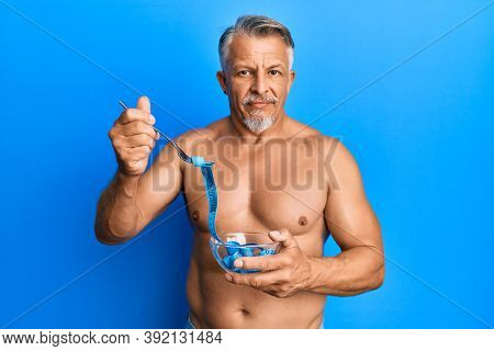 Middle age grey-haired man shirtless eating measure meter doing diet for weight loss relaxed with serious expression on face. simple and natural looking at the camera. 