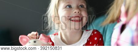 Portrait Of Smiling Cheerful Kid Putting Coin In Piggy Bank And Looking At Mother. Happy Joyful Chil