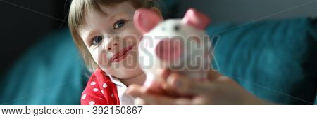 Portrait Of Lovely Little Kid Holding Pink Piggy Bank. Smiling Light Haired Girl Wearing Red Dress I