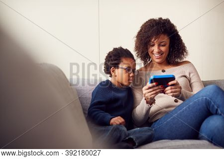 Mother And Son Sitting On Sofa At Home Playing Computer Game Together On Hand Held Device At Home