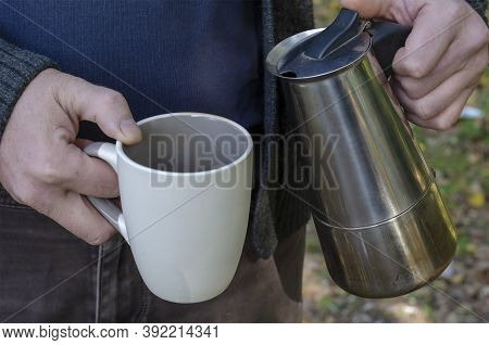 Man Pours Brewed Coffee Into Cup From An Outdoor Geyser Coffee Maker. An Adult Man's Hands Hold Cera