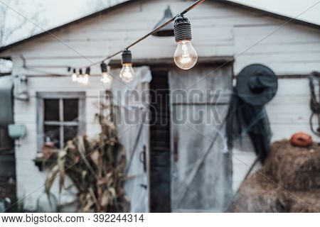 Light Bulbs On The Garland Near Old Scary Decorated Barn With Hay, Haystacks. Celebration Of Hallowe