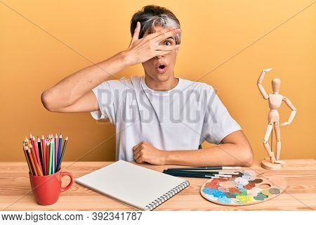 Young hispanic man artist sitting at the studio peeking in shock covering face and eyes with hand, looking through fingers afraid 