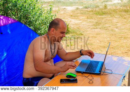 Man Using His Laptop Sitting At A Folding Picnic Table In A Camping. Working While Traveling. Freela