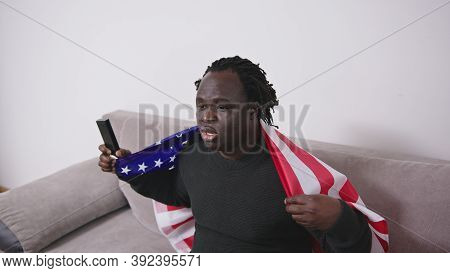 African American Black Man With Usa Flag Over The Shoulders Watching Sport Game On Tv On His Couch. 