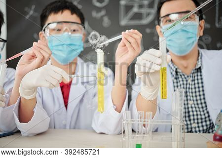 Excited School Students In Protective Masks And Goggles Adding Drops Of Reagent In Test Tube And Obs