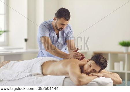 Serious Masseur Doing Relaxing Back Massage For Young Man Using Tapotement Technique
