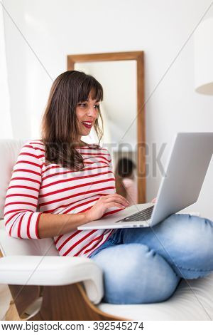 Successful young business woman sitting in a designer chair, working from home. Portrait of happy entrepreneur working on a laptop computer. Doing business remotely from the comfort of her home.