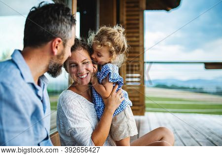 Family With Small Daughter Sitting On Patio Of Wooden Cabin, Holiday In Nature Concept.