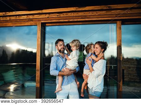 Family With Small Children Standing By Wooden Cabin, Holiday In Nature Concept.