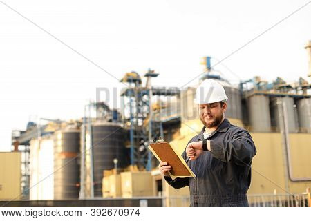 Caucasian Supervisor Looking At Watch And Holding Papers Notes Near Factory Outside.