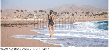 A Cheerful Woman In A Black Piecework Swimsuit Runs By The Sea .
