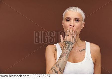 Manlike woman in white vest covering mouth by hand and showing arm with tattooes while sending air kiss or expressing astonishment