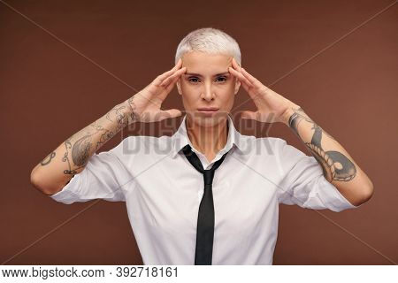 Manlike strict blond female with tattoos all over her arms keeping hands on temples while looking at you in isolation on brown background