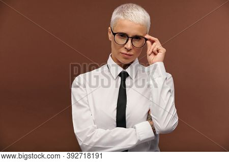 Young serious manlike woman in formalwear touching her eyeglasses while slightly bending aside and looking at you against brown background
