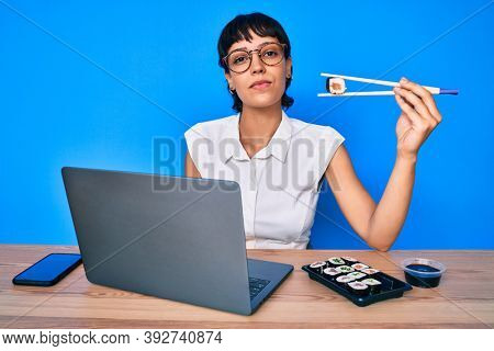 Beautiful brunettte woman working at the office eating sushi relaxed with serious expression on face. simple and natural looking at the camera. 