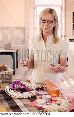 Happy casual caucasian blonde mid adult woman making valentine day wreath at home. Smiling, wearing glasses, looking at camera, standing at table, do it yourself, gift, present.