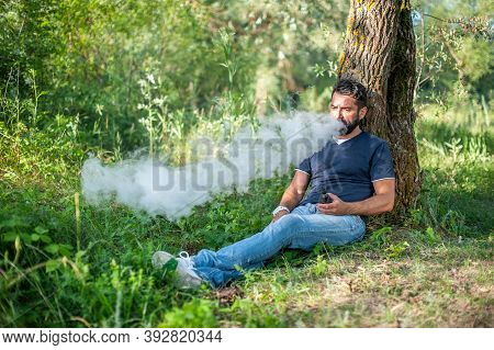 Stylish Brutal Vaper Smoking An Electronic Smoke Device On The Forest Ground. Alternative To Smoking