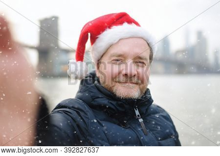 Middle Aged Man Tourist In Santa Claus Hat Takes Selfie In Front Of Stunning Views Of Brooklyn Bridg