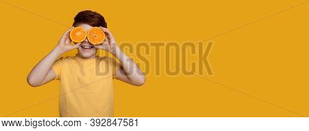 Small Caucasian Ginger Boy Is Covering His Eyes With Oranges Posing In A T-shirt On A Yellow Studio 