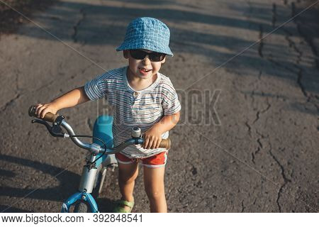 Caucasian Boy With Blue Hat And Sunglasses Smiling At Camera While Holding The Bike On The Road