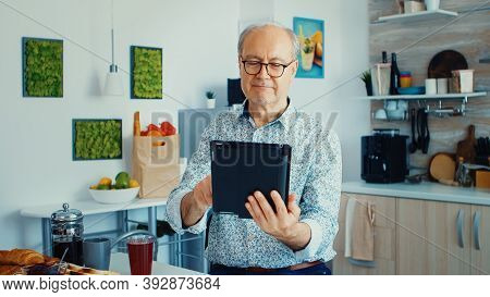 Happy Older Man In Kitchen During Breakfat Using Tablet Pc. Elderly Person With Tablet Portable Pad 