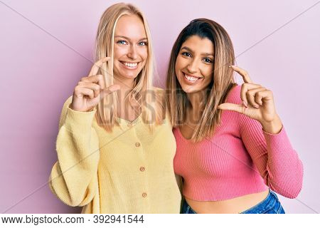 Two friends standing together over pink background smiling and confident gesturing with hand doing small size sign with fingers looking and the camera. measure concept. 