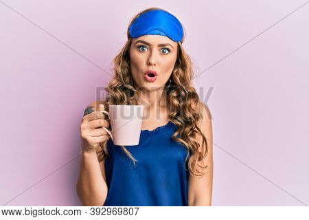 Young blonde girl wearing sleep mask and pyjama drinking coffee scared and amazed with open mouth for surprise, disbelief face 