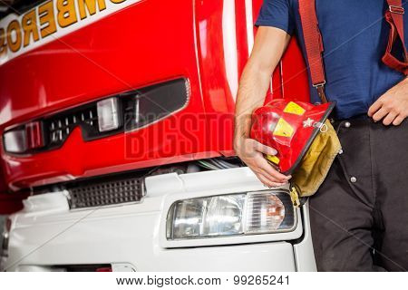 Midsection of male firefighter holding red helmet while leaning on firetruck at station