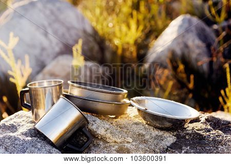 Set of camping cookware pot, bowl, pan, mug and utensils outdoor in the morning sunlight at a remote mountain wilderness campsite