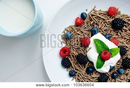 Muesli With Berries In White Dish On Blank White Table With Blue Cup Filled With Milk Top View