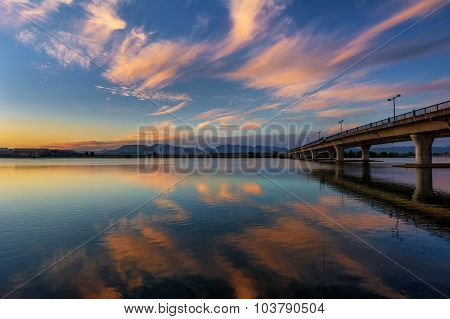Playful clouds, pastel-coloured sky and water, concrete bridge, and mountains